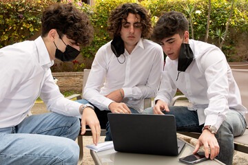 A group of young students are sitting at a table, working at a laptop, they are wearing masks ,
