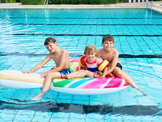 Three kids, two boys and toddler girl splash in an outdoors swimming pool in summer. Happy children, brothers and sister playing on huge inflatable gum lollipop enjoying sunny weather in public pool.