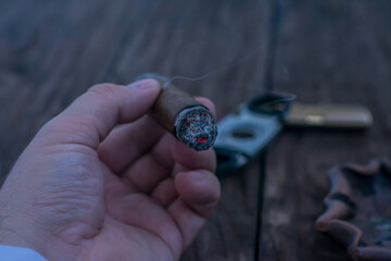 A man's hand holds a smoldering cigar over a wooden table against the background of a guillotine, a...
