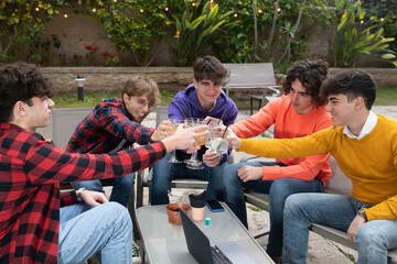 A group of students 18 y old are sitting in a bar, cheering and toasting with drinks in hands, celebrating a victoring or a success. A laptop and a gel bottle is on the table