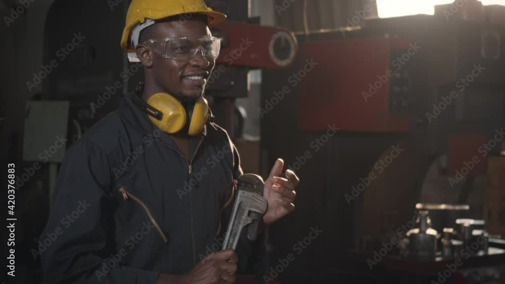 Wall mural Portrait American industrial black young worker man smiling with helmet and ear protection in front machine, Engineer standing holding wrench on his hand at work in industry factory. slow motion