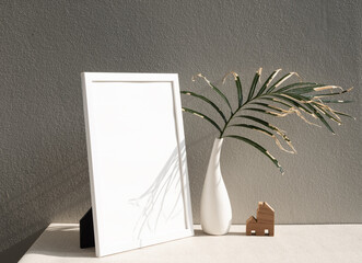 Mock uo poster frame and dry palm leaves  botanical tropical house plant in beautiful white ceramic vase andhouse model on earth tone  table and cement  wall background