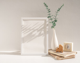 Mock up white wood poster frame,Eucalyptus  dried leaf in vase,clock, books, house model on beige table and wall,modern stylish home decor interior with long shadow