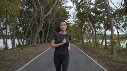 asian athlete woman in black sportswear and wireless headphone running in urban city trees park during sunset and bad cloudy weather. female jogging outdoor in evening. healthy workout.