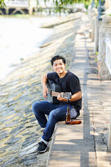 Portrait of Asian young man smiling at camera while playing guitar on the street outdoors