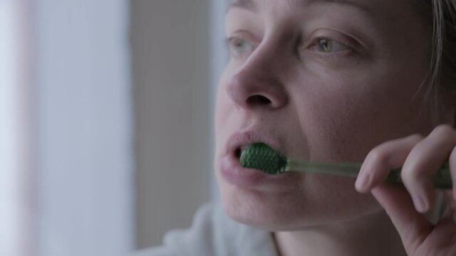 A woman brushes her teeth while holding a toothbrush in her left hand, standing by the window