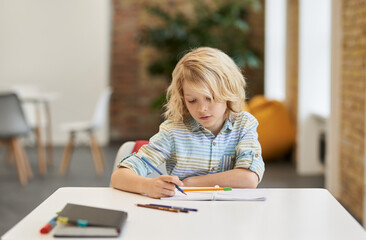 Doing assignment. Smart little school boy writing in his notebook while sitting at the desk in classroom