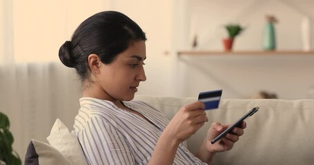 Side view relaxed smiling millennial indian woman entering payment information from credit bank card in mobile shopping app, purchasing goods or services in internet store or booking tickets. - Powered by Adobe