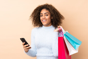 Young African American woman isolated on beige background holding shopping bags and writing a message with her cell phone to a friend