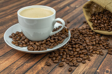 A cup of creamy coffee with roasted coffee beans next to it, wooden background