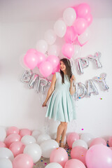 Smiling brunette woman in mint dress holding a lot of baloons. Birthday decorations with white and pink color balloons and confetti for party on a white wall. Happy birthday anniversary. Girls party.