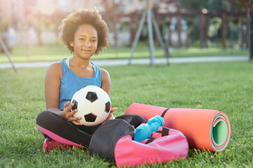 Naklejka premium Cheerful little girl with soccer ball sitting on grass outdoors