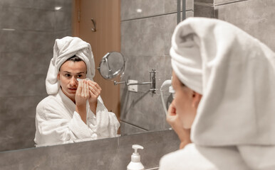 A young woman in a robe and with a towel on her head is putting patches under her eyes.