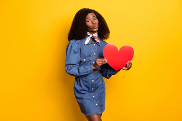 Portrait of charming dark skin girl kiss lips hands hold red paper heart postcard isolated on yellow color background