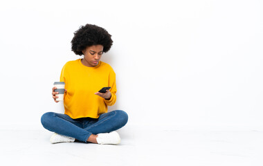 Young African American woman sitting on the floor holding coffee to take away and a mobile