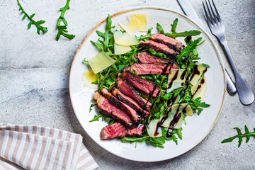 Tuinposter Beef steak tagliata with arugula and parmesan on gray plate, gray background, top view. Italian cuisine concept. © vaaseenaa