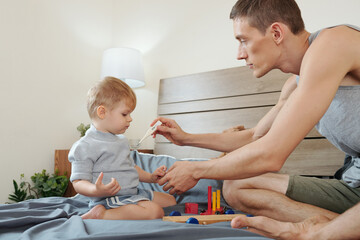 Little boy sitting on bed and playing while his father measuring his temperature during illness