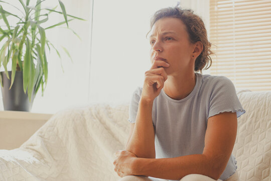 Pensive thoughtful young woman thinking pondering sitting on sofa at home looking in distance. Thinking regret about mistake, suffering from depression, emotional breakdown, psychological problem