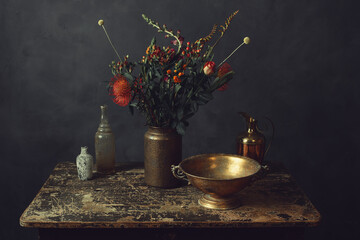 Old worn wooden table topped with a vase with flowers, a bronze bowl, pitcher and two glass bottles.