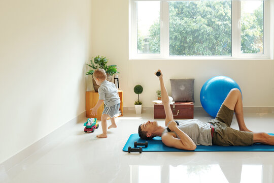 Young man lying on exercise mat and raising dumbbells while his son playing in the room