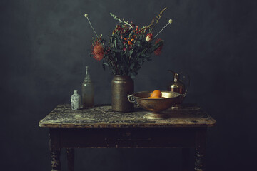 Old worn wooden table topped with a vase with flowers, a bronze bowl with oranges, a pitcher and...
