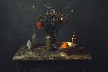 Old worn wooden table topped with a vase with flowers, a bronze bowl with oranges, a pitcher and...