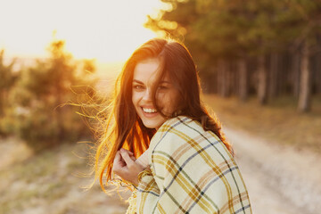 woman with a blanket on her shoulders laughs the sunset in the background and conifers on the side