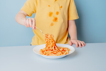 Cute boy eating spaghetti bolognese at home. concept of dirty stains on clothes. isolated on blue...