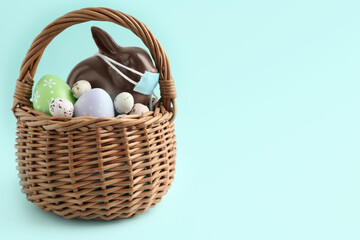 Chocolate bunny with protective mask and eggs in basket on light blue background, space for text. Easter holiday during COVID-19 quarantine