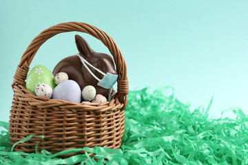 Chocolate bunny with protective mask and eggs in basket on light blue background, space for text....