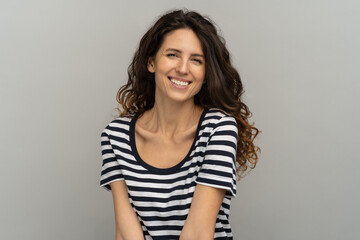 Fototapeta premium Portrait of European happy woman with curly hair and toothy smile, looking at camera. Pretty girl wear stripped t-shirt laughing, having fun standing isolated on studio grey background with copy space