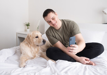A young man holds a mobile phone while sitting on a bed with a dog and caresses his pet. Portrait of male owner spending time at home in bedroom scratching golden retriever