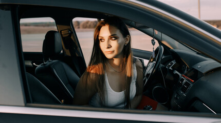 Obraz na płótnie Canvas Beautiful young girl driving a car in the evening in the sunset sun on an empty parking lot