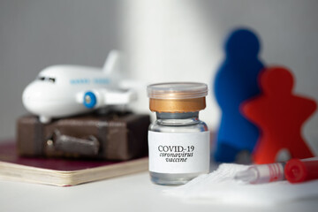 Vial  with COVID-19 vaccine and syringe on a sterile gauze. Red and blue wooden figurine, passenger aircraft, luggage and travel pass in the background.