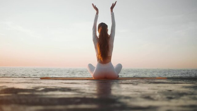 A back-view of a young redhead woman athlete wearing white sportsuit is doing yoga sitting outside near the sea in the morning