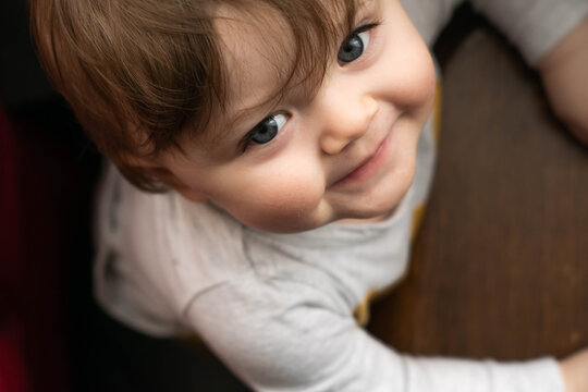 Caucasian Baby Boy With Long Brown Hair And Blue Eyes Smiling And Looking Up Straight To The Camera Above. Happy Baby Top View.