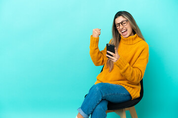 Young caucasian woman sitting on a chair isolated on blue background with phone in victory position