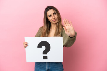 Young caucasian woman isolated on pink background holding a placard with question mark symbol and doing stop sign
