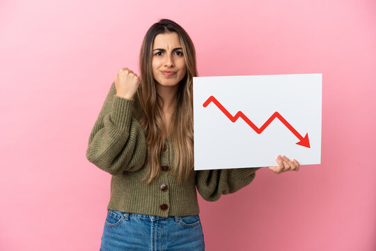 Young caucasian woman isolated on pink background holding a sign with a decreasing statistics arrow symbol and angry