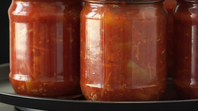 Glass jars with canned lecho rotates on tray. Canned food with bell peppers, tomatoes, spices and seasoning