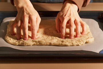 Woman preparing focaccia. Women's hands crumpling raw dough on metal tray, pressing fingers and pushing holes in recesses in raw dough. Homemade, cooking at home