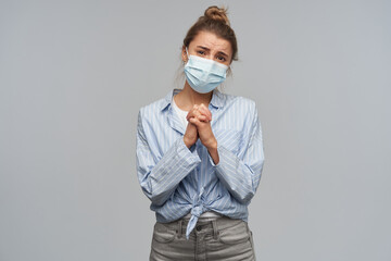 Begging woman, asking girl with blond hair gathered in bun. Wearing striped knotted shirt and protective face mask. Keeps palms pressed together. Watching at the camera, isolated over grey background