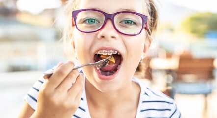 Young girl wearing glasses and braces puts food on fork into her mouth - obrazy, fototapety, plakaty