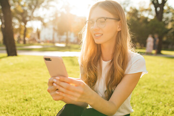 young woman in glasses sitting in the park on the lawn using a mobile phone
