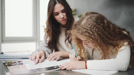 Pretty mother helps her daughter doing her homework sitting at the table at home