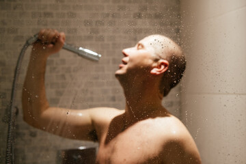Positive young man having fun in bathroom, is singing with shower as a microphone. Personal hygiene concept. Defocused and blurred photo.