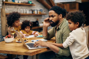 Pensive black father working on computer while being with his family at home.