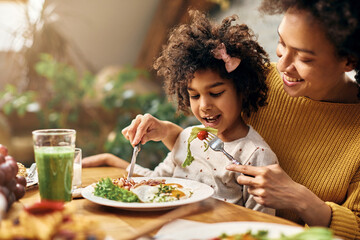 Happy black mother feeding daughter while she is sitting on her lap at dining table.