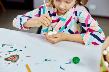 Happy little girl painting, drawing with brush eggs at home.