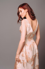 Ginger young woman in golden peach dress with floral print. Calm studio portrait of young lady in long sleeveless evening gown on grey background.